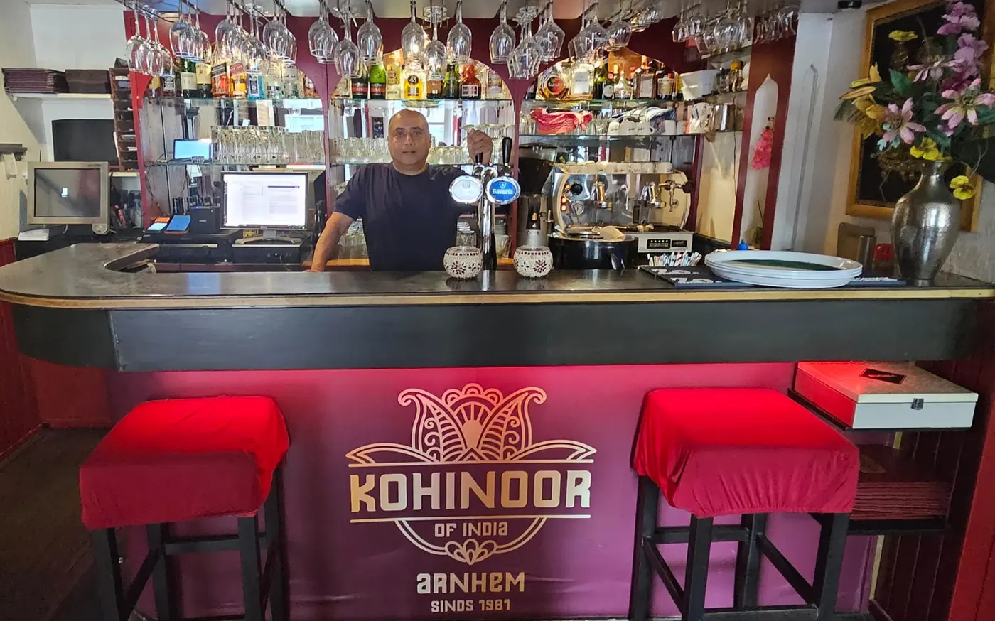 De bar van Kohinoor of India met rode barkrukken en het logo, met de eigenaar achter de bar