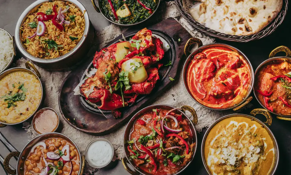 Een tafel vol Indiase gerechten in koperen karahi's: curry's, biryani, tandoori kip en naan, op een donkere leistenen tafel